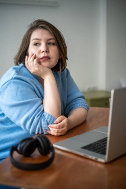 Büyük beden genç bir kadın başını elinin üzerine koyup dikkatlice uzaklığa bakıyor. Dizüstü bilgisayar ve kulaklıklarla masada oturuyordu..
