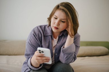 Yatakta oturan bir kadın, düşünceli bir ifadeyle akıllı telefona bakıyor. Günlük yaşam, teknoloji kullanımı ve dijital iletişim.