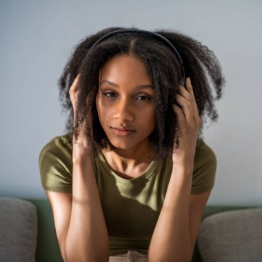 Genç, kıvırcık saçlı, kulaklık takan, koltukta oturan ve kameraya bakan Afro-Amerikan kadın..