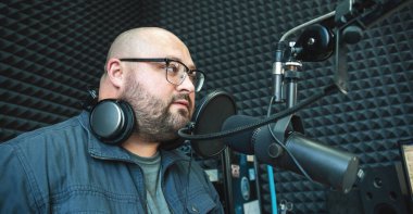 Ses geçirmez kayıt stüdyosunda gözlüklü bir adam, mikrofona odaklanmış, sese ya da podcast kaydına hazır. Kulaklık ve akustik köpük duvarlı profesyonel ses düzeneği.