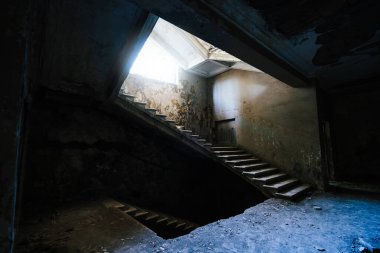 Eski harap binanın içindeki terk edilmiş merdiven. Kırık tavandan güneş ışığı geliyor. Atmosferik bozulma, duvarların soyulması ve gizemli ışıklandırma unutulmuş bir yerde ürkütücü bir ruh hali yaratıyor..