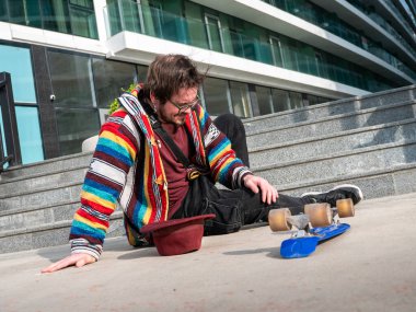 Skateboarder sitting on pavement after fall, wearing colorful striped jacket, urban background with stairs and modern building facade, skateboard in front. clipart