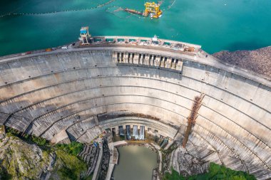 Üzerinde su boşaltım kanalları ve inşaat vinci olan devasa beton baraj. Hidroelektrik yapı ve turkuaz rezervuarın kayalık araziyle çevrili havadan görüntüsü. Yenilenebilir enerji