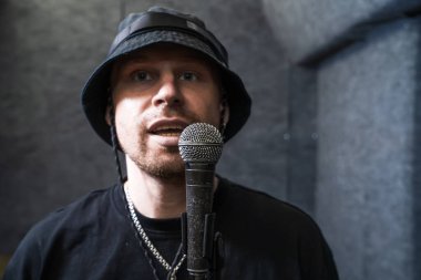 Siyah şapka takan rapçi, müzik stüdyosunda mikrofona şarkı söylüyor, canlı vokal performansı ve hip-hop havasını yakalıyor.. 