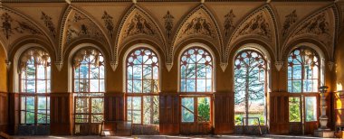 Gotik tarzda terk edilmiş bina içi uzun kemerli pencereler, karmaşık tavan detayları ve çürümüş ahşap paneller. Kırık pencerelerden geçen güneş ışığı unutulmuş olanları vurgular.