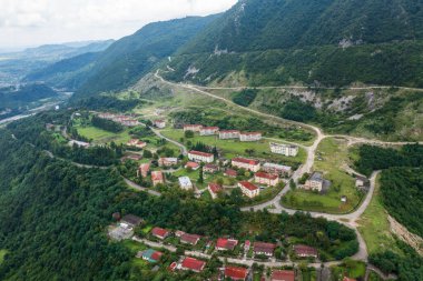 Yeşil tepelerle çevrili birden fazla apartmanı olan terk edilmiş bir dağ yerleşiminin havadan görüntüsü. Virajlı yollar yapıları birbirine bağlıyor, insan varlığı ile insan varlığı arasında bir kontrast oluşturuyor.