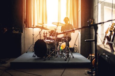 Davulcu müzik stüdyosunda çalacak, ılık güneş ışığı ile prova oturumu, grup çalışması, indie rock vibe, ses üretimi, yaratıcılık.