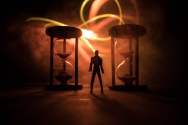 Zaman kavramı. Kum saatleri arasında duran, dumanlı ve karanlık arka planda ışıkları olan bir adamın silueti. Gerçeküstü dekore edilmiş resim