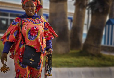 Malaga yakınlarındaki Torrox Costa plajında kolye satan renkli Afrikalı bir kadın. Büyük çiçek desenli kırmızı bir elbise giyiyor..