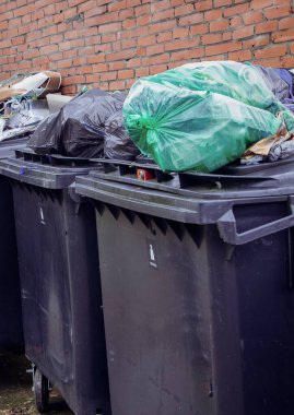 Üstünde yeşil ve siyah plastik poşetler olan şişeler, teneke kutular ve diğer çeşitli eşyalar içeren aşırı dolu bir çöp konteynırı. Olay yeri uygun olmayan atık imhası vurguluyor.