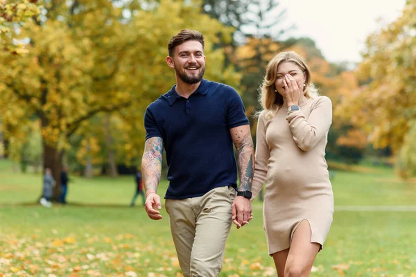 时尚的年轻孕妇和她英俊的丈夫正在秋天的公园里散步 — 图库照片