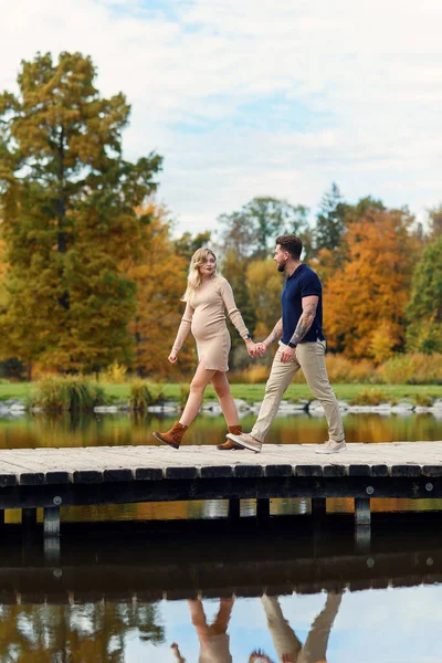 秋日公园 一位时尚的年轻孕妇和她英俊的丈夫手牵手走在桥上 穿过湖面 — 图库照片