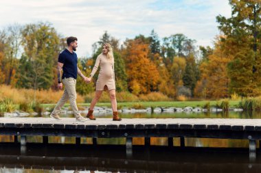 Havalı genç hamile kadın ve yakışıklı kocası sonbahar parkındaki gölde ahşap köprüde el ele yürüyorlar..