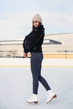 Şık ceketli çekici genç bir kadın Noel ışıklarıyla süslenmiş açık havada buz pateni pistinde vakit geçiriyor. Neşeli genç bayan Noel buz pateni pistinde kayıyor..