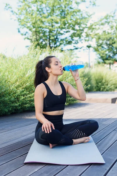 Sporty young woman drinks energy drink during warm up fitness exercises with yoga mat at the park outdoor.