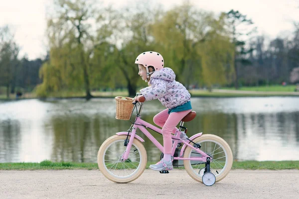 大きな湖の近くの日当たりの良い春の公園で安全なヘルメットで自転車に乗っている女の子 安全なヘルメットで自転車にバランスをとることを学ぶ未就学児 ストックフォト