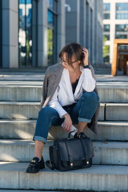 Takım toplantısından sonra iş çantalı güzel bir iş kadını modern ofis binasının yanındaki merdivenlerde düşünceli bir şekilde oturuyor. Başarılı kadın yönetici kavramı.