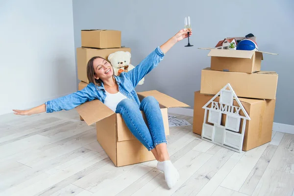 Pretty beautiful girl raised her hands up while moving to the new home. Funny woman with glass of champagne sits in the cardboard box.