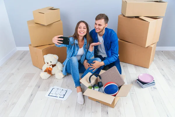 Young attractive couple taking selfies in their new home while sitting among cardboard boxes. Happy attractive young couple is moving, making a selfie, cuddling and smiling.