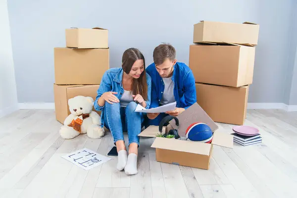 Happy caucasian couple have discussion about design of their new home sitting on the wooden floor between cardboard boxes. Beautiful stylish couple in love planning design for their new flat. Top view