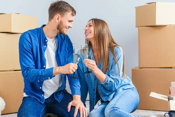 Young couple taking a break on moving day, sitting on floor in new apartment celebrating with champagne surrounded by cardboard boxes.
