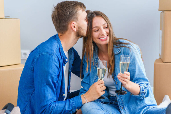 Young couple taking a break on moving day, sitting on floor in new apartment celebrating with champagne surrounded by cardboard boxes.