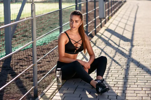 A beautiful girl in black sportswear listening to music with wireless headphones while training on an outdoors sports ground. Sports concept.
