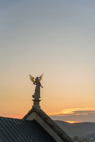 Back view of stone sculpture of an angel with golden wings standing on the rooftop of a baroque temple at sunset.