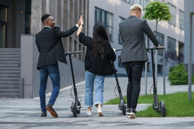 İş projesini tartışan çok ırklı ofis arkadaşları elektrikli scooterlarla ofis binasının yakınına gidiyorlar.