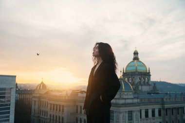 Siyah ceketli çekici kıvırcık esmer, büyülü bir günbatımında büyük tarihi bir binanın arka planında duruyor. Yumuşak güneş ışığı teninde.