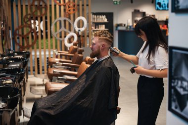 Berber dükkanında oturan kızıl saçlı sakallı adamın ve kadın berberin misafir için saç kesiminin yan görüntüsü.