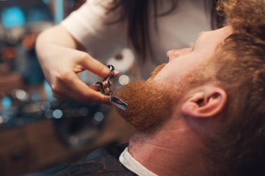 Berber dükkanında oturan kızıl saçlı sakallı adamı ve makasıyla sakal düzeltme yapan kadın berberi yan görüyorum..