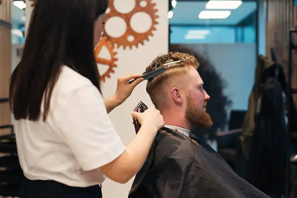 stock image Side view of red-haired bearded man sitting in barbershop and female barber making hairdo to guest