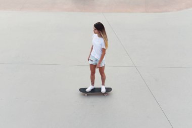 Beyaz tişörtlü ve kot şortlu kaykaycı kız modern şehir kaykay parkında kaykay sürüyor. Modern hipster yaşam tarzı.