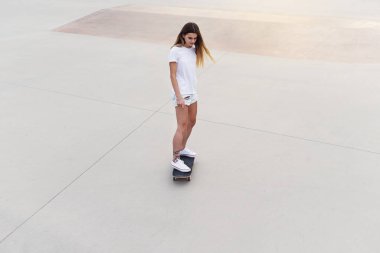 Beyaz tişörtlü ve kot şortlu kaykaycı kız modern şehir kaykay parkında kaykay sürüyor. Modern hipster yaşam tarzı.