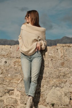 The girl stands in front of a stone wall and looks to the left