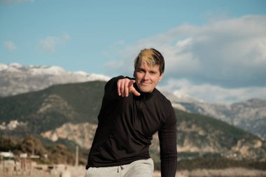 Genç bir adam kameranın önünde duruyor ve işaret parmağını lense doğrultuyor.