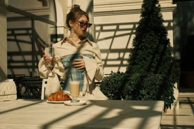 Bir kız açık bir kafede masada oturuyor, elinde bir bardak su tutuyor.