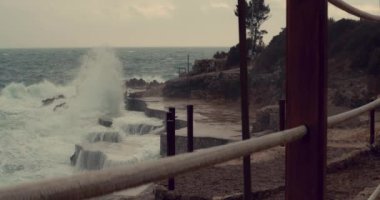 Karadağ 'da bir plajda büyük dalgalar siyah kayalara çarpıyor. Akdeniz 'in sinematik videosu, kayalık bir sahil şeridi boyunca kabaca sıçrıyor, el kamerasıyla ip çitten geçiyor.