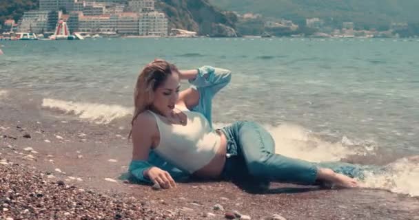 湿ったデニムの服を着た若い女性が海岸波の中でカメラのポーズをとっている 背景には山や街が見える スローモーション映画の映像 映画的な眺め — ストック動画