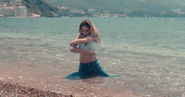 濡れたデニムの若い女性が水の中に座って 彼女の濡れた白い上を削除します スローモーション映画の映像 映画的な眺め — ストック動画