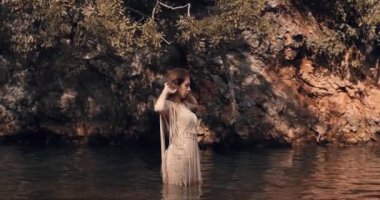 Genç bir kız beline kadar suya batmış, saçını düzleştiriyor ve kameraya poz veriyor. Arka planda, kayanın üzerindeki sudan gelen parıltı görülebilir. Boho. Islak Bakış tarzı. Sinematik görünüm