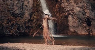 Küçük bir gölün kıyısında beyaz şapkalı genç bir kız ekseni etrafında dönüyor. Arka planda bir şelale görülebilir. Boho. Sinematik görünüm