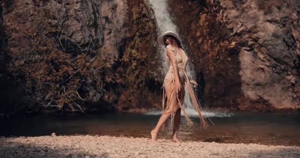 小さな湖の岸に白い帽子をかぶった若い女の子が太陽の下で彼女のドレスで遊んでいます 背景には滝が見える シネマティック ビュー — ストック動画