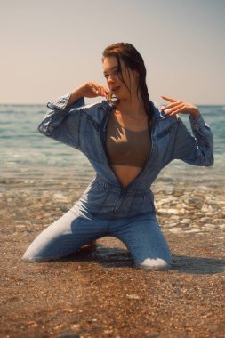 Düğmeleri iliklenmemiş ıslak kotlu bir kız dalgaların arasında oturur ve kameraya poz verir. Kollarını kaldırdı. Onun bakışı kamera lensine doğru yönelir. Omuzlarında ıslak saçlar var. Islak görünüm tarzı