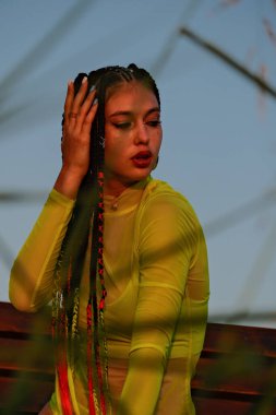 Uzun saçlı ve sarı gömlekli bir kadın bankta oturuyor. Görüntü, kadının renkli kıyafetleri ve güneşli günüyle neşeli ve neşeli bir ruh haline sahip. Çimlerin arasında fotoğraf..