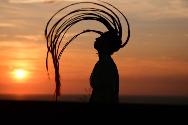 Uzun saçlı bir kadın, canlı bir günbatımına karşı siluetlenir ve büyüleyici bir görüntü yaratır. Kısmen belirsiz figür esrarengiz bir güzelliğe işaret ediyor. Yumuşak odak.