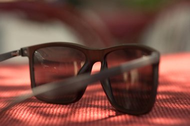 Kırmızı bir masada oturan bir güneş gözlüğü. Güneş gözlükleri siyah ve plastik çerçeveleri var. Güneş gözlükleri sıcacık bir yüzeye yerleştirildiği için görüntüde sıcak ve davetkar bir hava var.