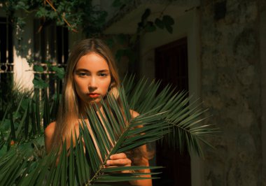 Elinde yapraklı yeşil bir bitkiyle bir binanın önünde duran bir kadın. O, siyah bir elbise giyiyor ve üzerinde kırmızı bir ruj var. Görüntünün doğal ve rahat bir havası var.