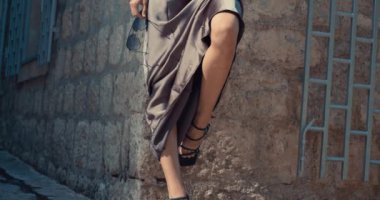 Tuğla duvarda siyah sandaletler giyen bir kadın var. Uzun bir elbise giyiyor ve bacak bacak üstüne atmış.